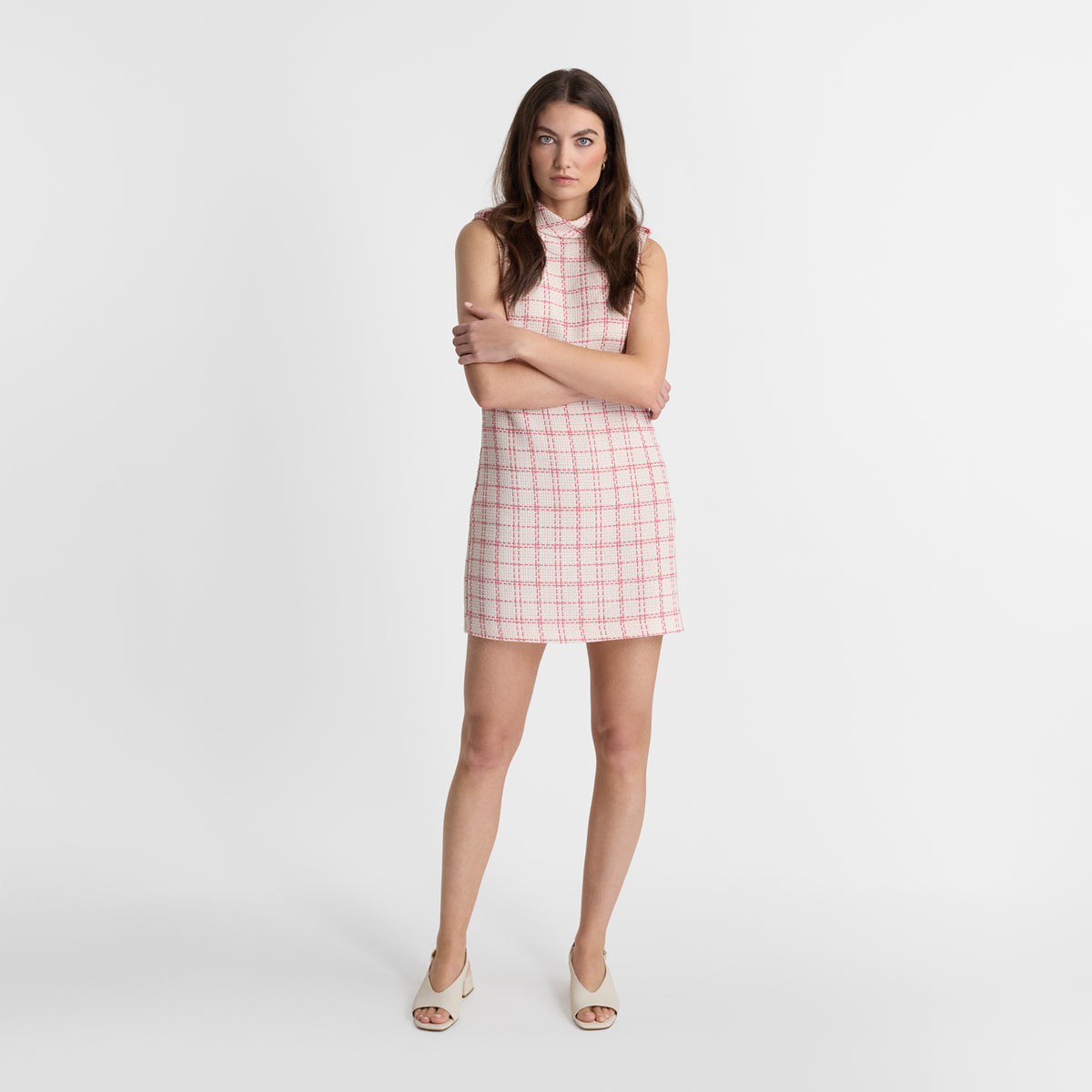 A woman stands against a light gray studio background wearing a sleeveless, high-neck mini dress with a pink and white plaid pattern and cream-colored heeled sandals.
