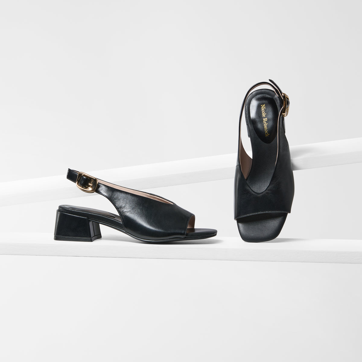 A pair of black leather slingback block-heel sandals with gold-tone buckle details, displayed on a clean white geometric platform.