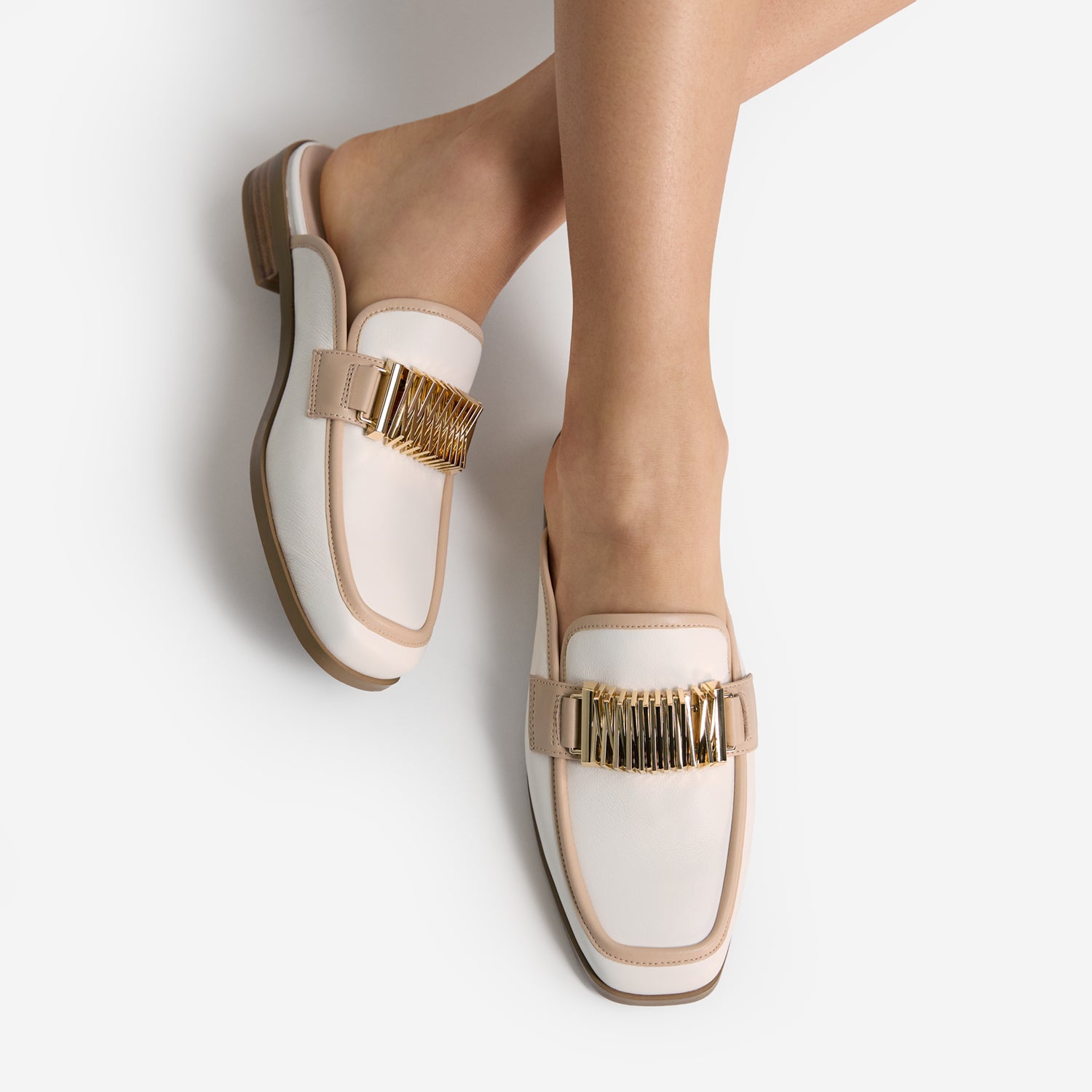 White mule loafers with gold embellishments worn by a person on a light grey background