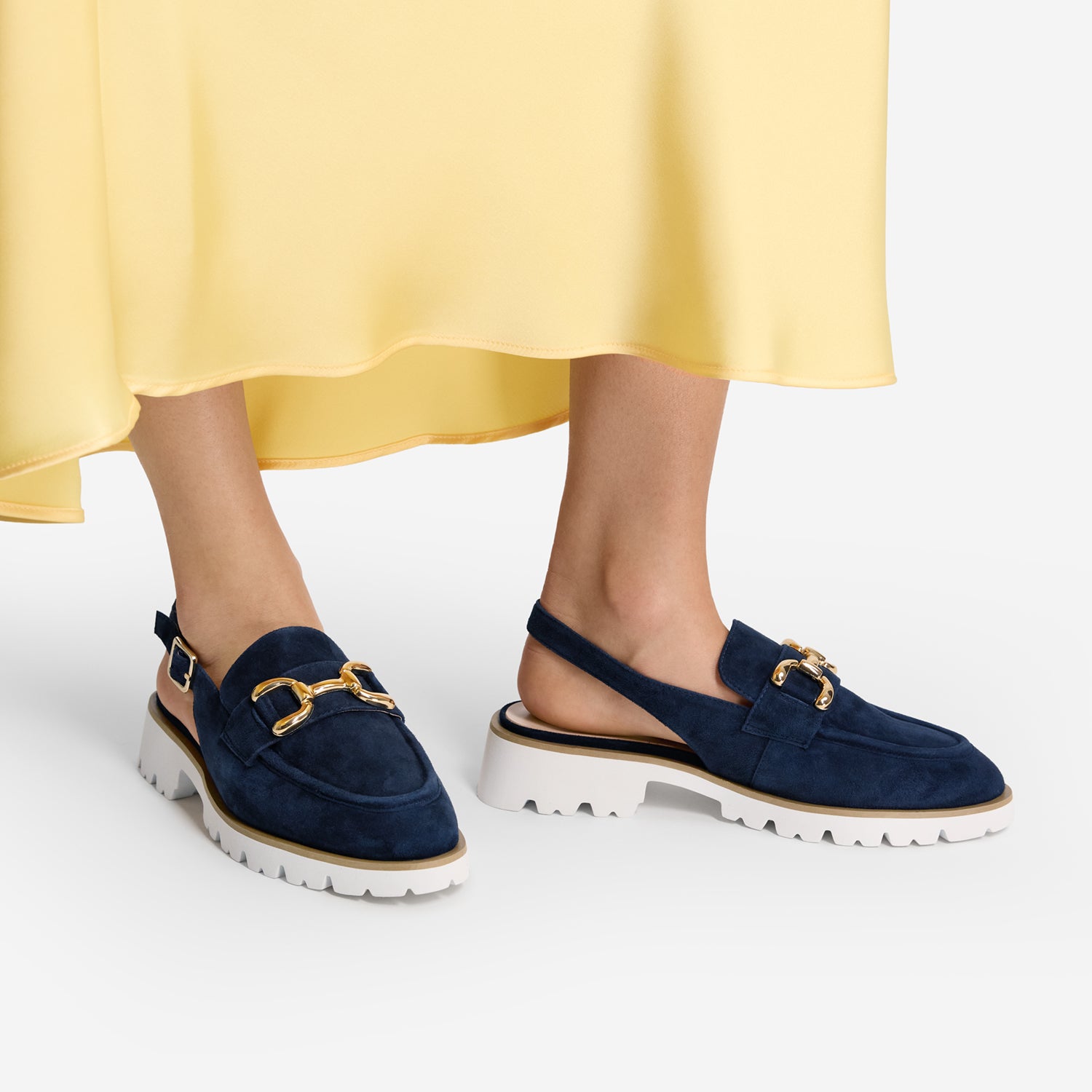 Navy loafers with gold accents worn with a yellow skirt on a white background
