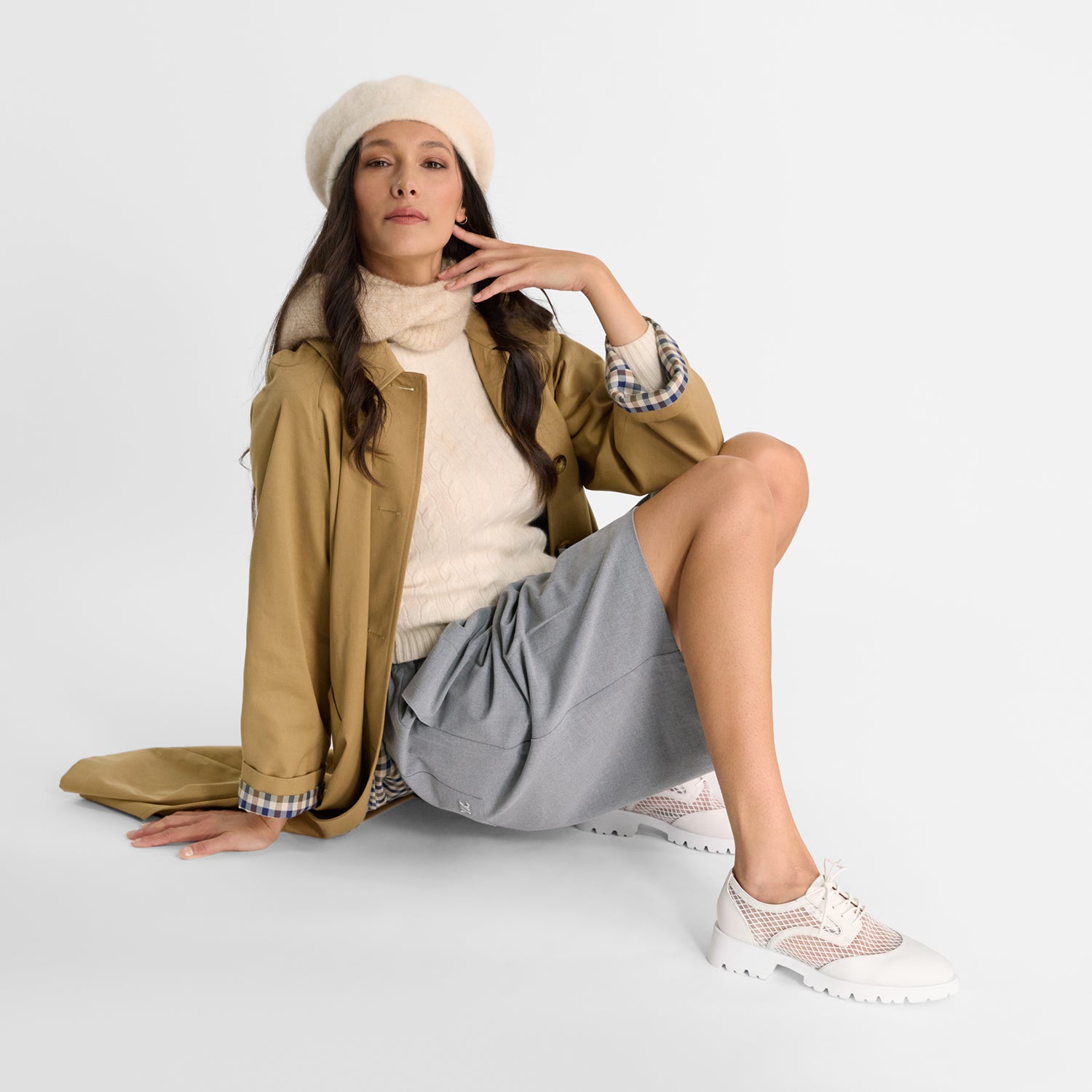 Woman sitting on a white surface wearing a beige coat, white sweater, gray skirt, and white cassidy lace-up flats.
