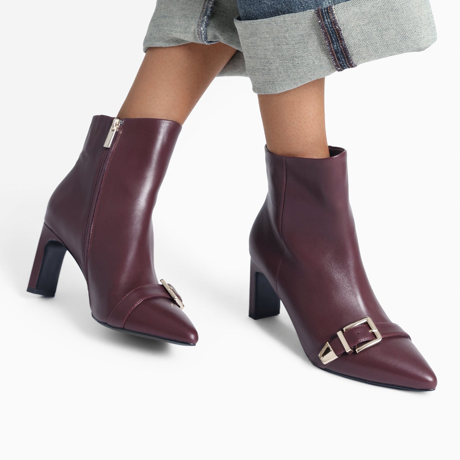 Close up of Rome Ankle Boots worn by women. The boots are oxblood colour with gold buckles over the pointed toes. The heel is a block rectangle with a slim side profile. Heel height is 7.5cm.