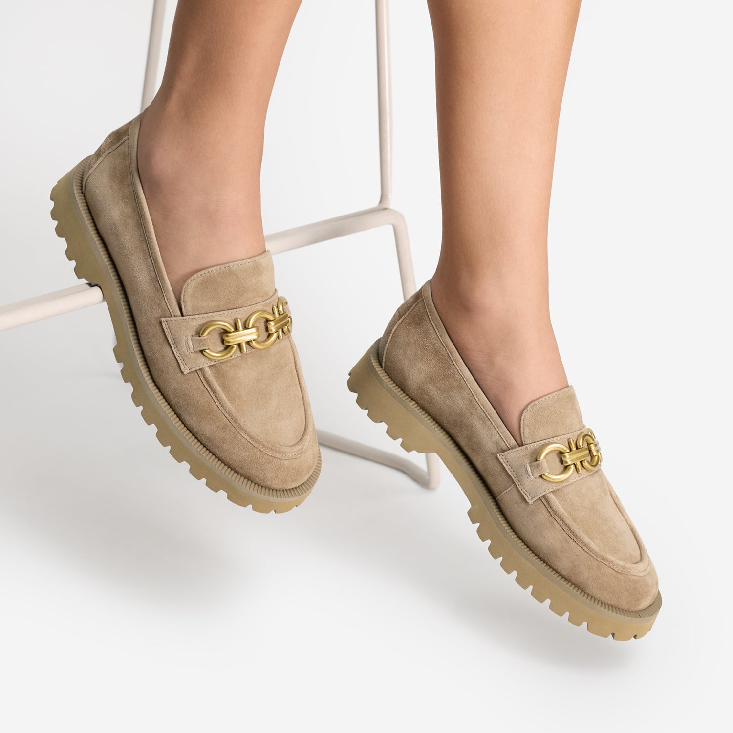 A pair of beige suede loafers with chunky soles and a gold-tone chain detail across the vamp, shown on a model's feet sitting on a white metal stool.