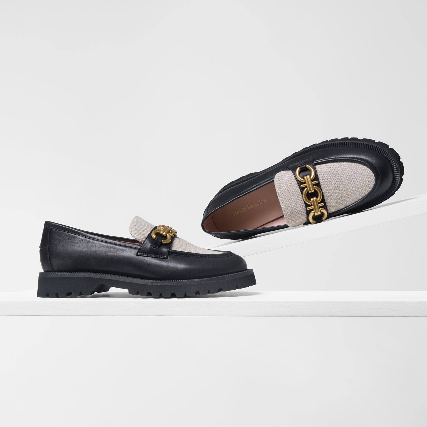 A pair of black leather loafers with a chunky EVA sole, featuring a light linen tongue across the vamp and a prominent gold-tone chain hardware on the strap. One shoe is shown from a side profile on a white platform, and the other is positioned above it showing the top and sole.