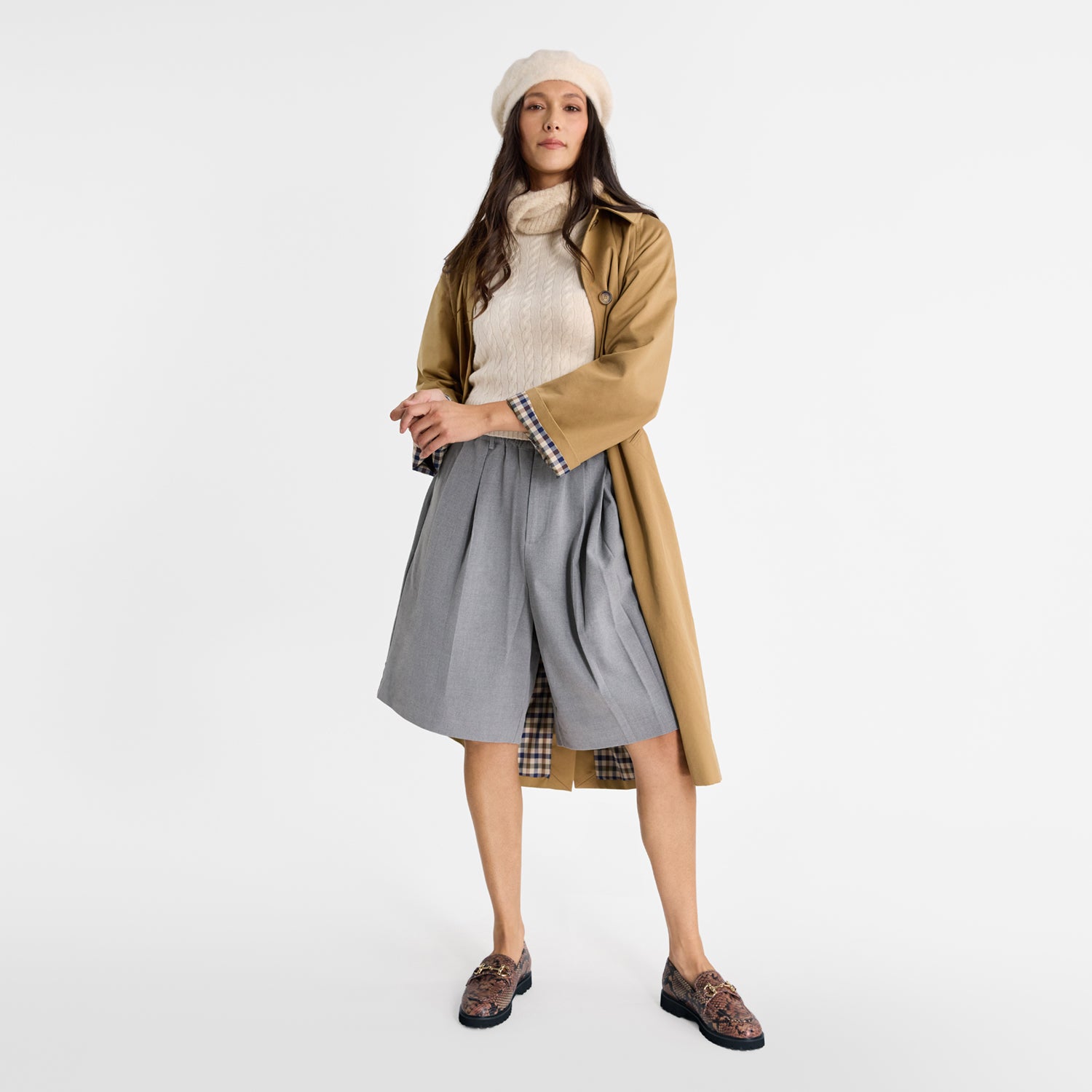 Woman wearing a beige coat, gray skirt, white sweater and brown loafers on a white background