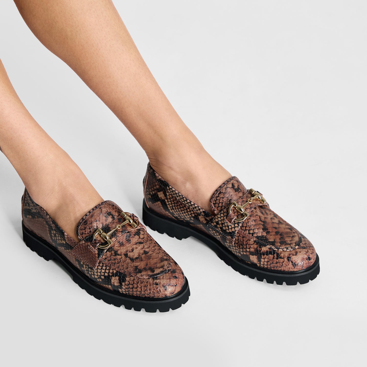 Snack patterned brown loafers worn by a person on a light gray background
