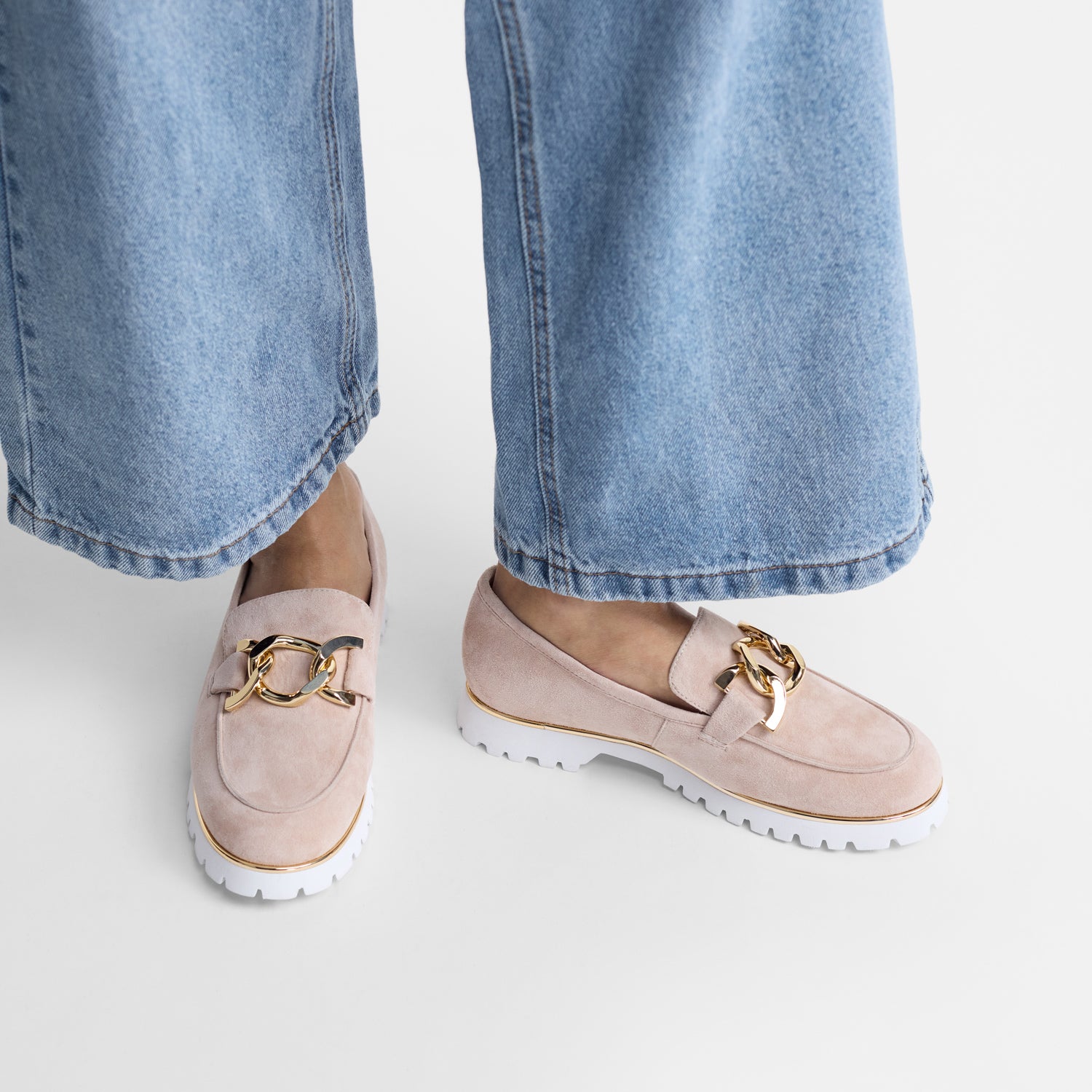 Nude suede loafers with gold accents worn with light blue jeans on a white background
