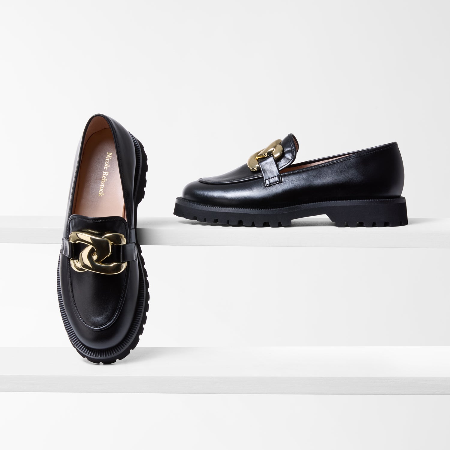 Black loafers with gold accents on a white background by Nicole Rebstock