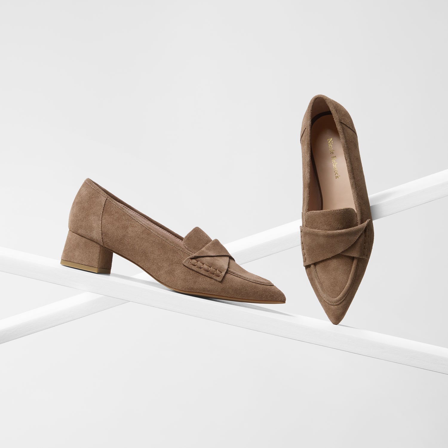 A pair of taupe-brown suede loafers with low block heels, shown on a clean white geometric display stand by Nicole Rebstock.