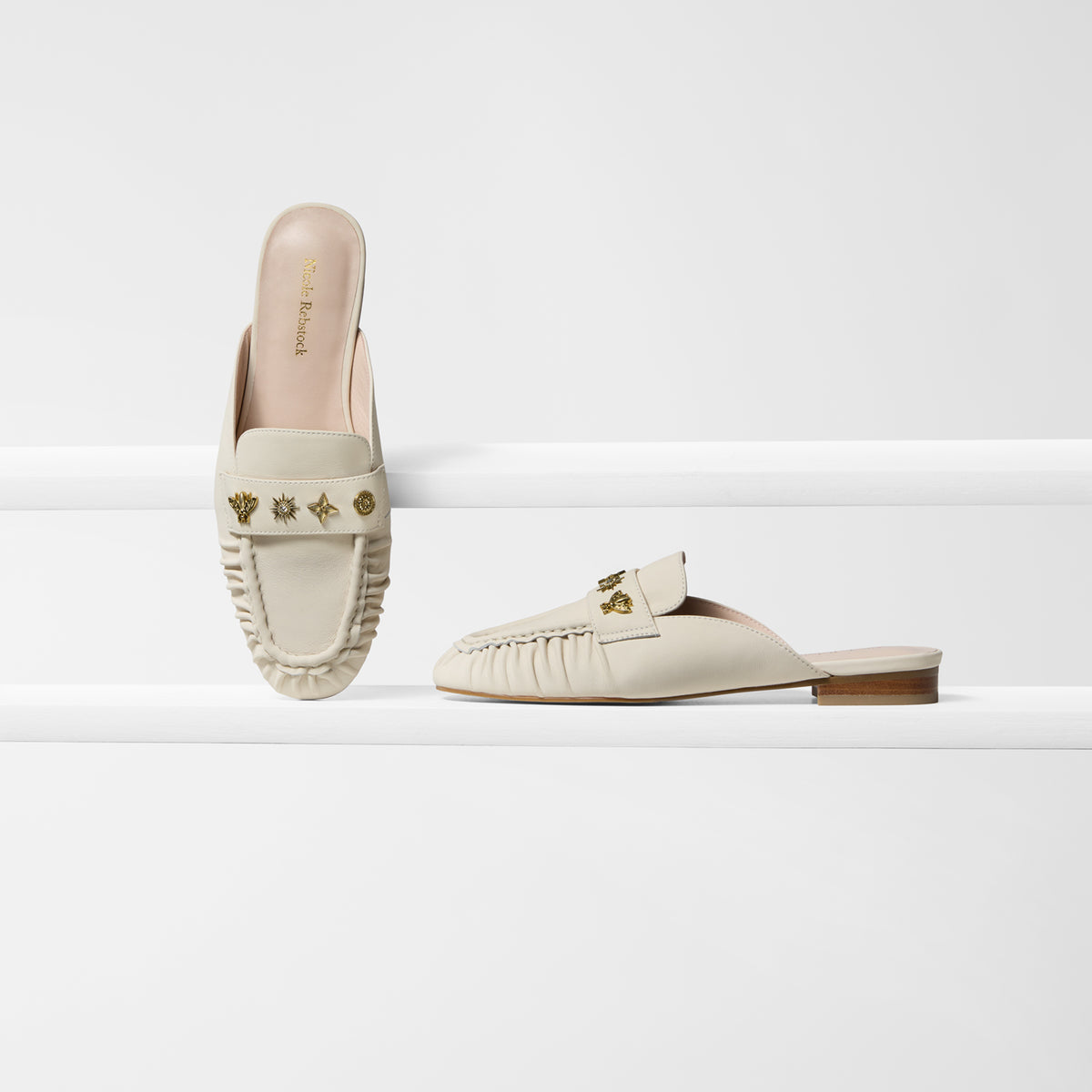 A pair of cream-colored leather slip-on mule loafers with gold-tone hardware and decorative stitching, displayed on a clean white geometric background.
