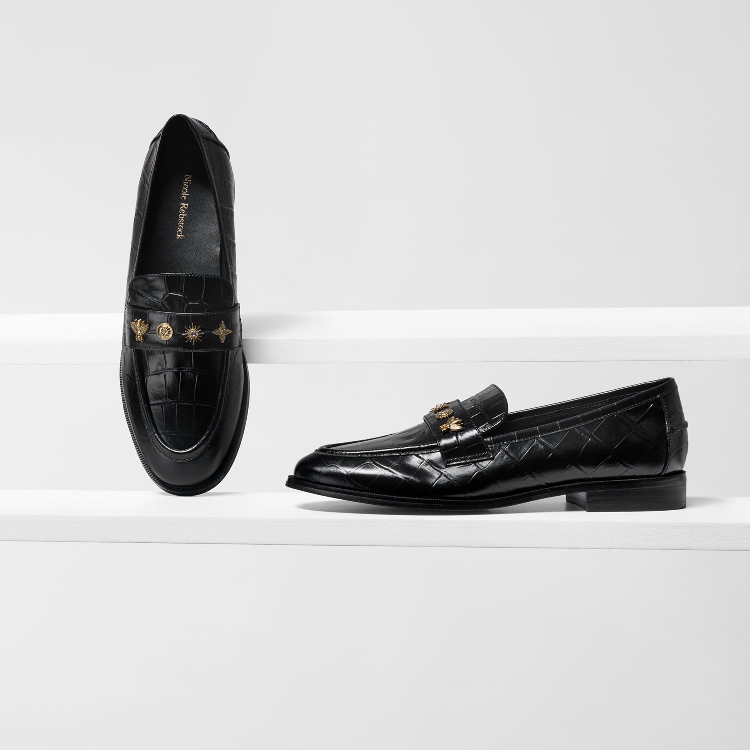 A pair of black leather loafers displayed on white steps against a white background. One shoe is shown from the front on its side, revealing gold-tone hardware and a crocodile-embossed texture, while the other is shown from the side.