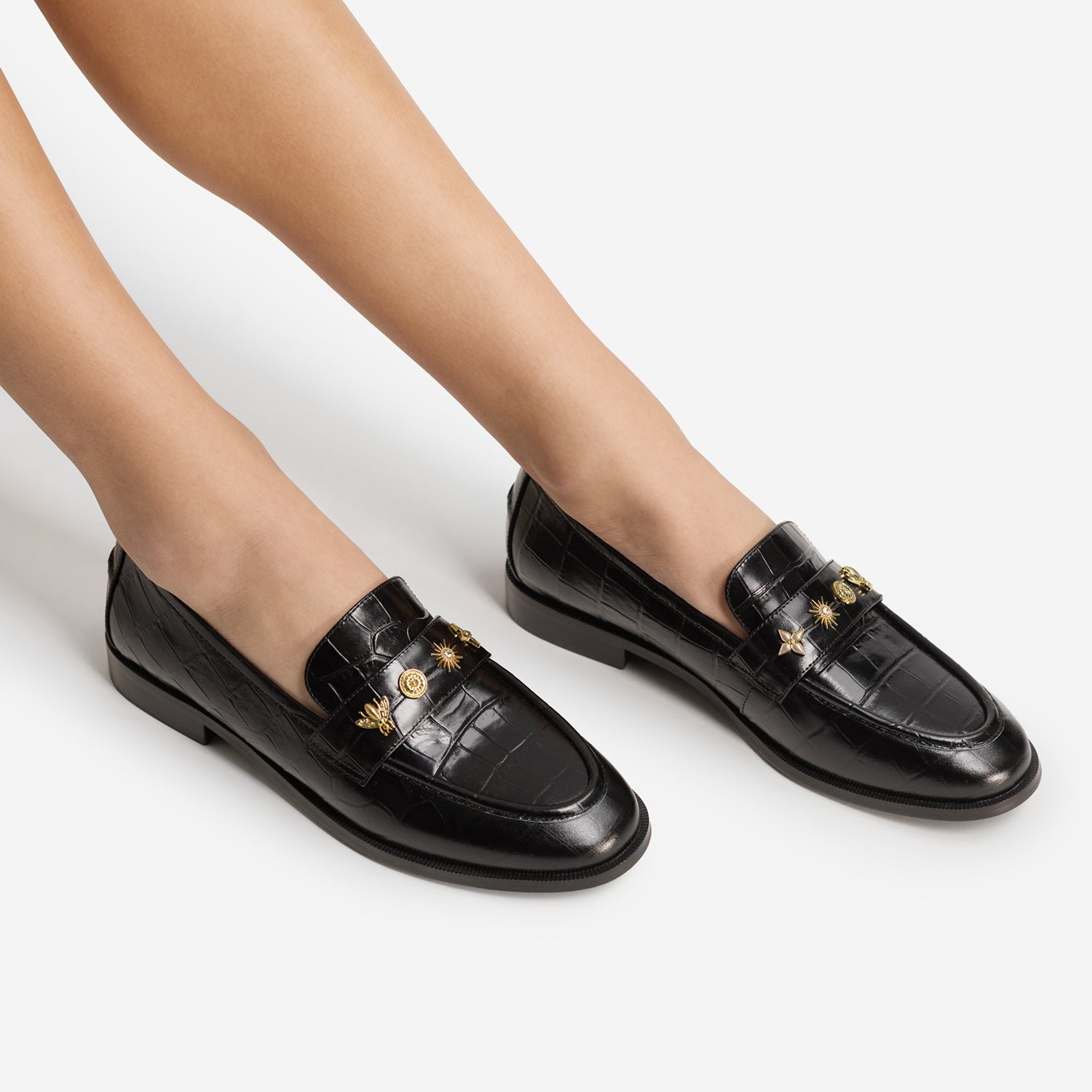 A pair of black crocodile-embossed leather penny loafers with gold-tone decorative hardware, shown on a model's feet against a light gray studio background.