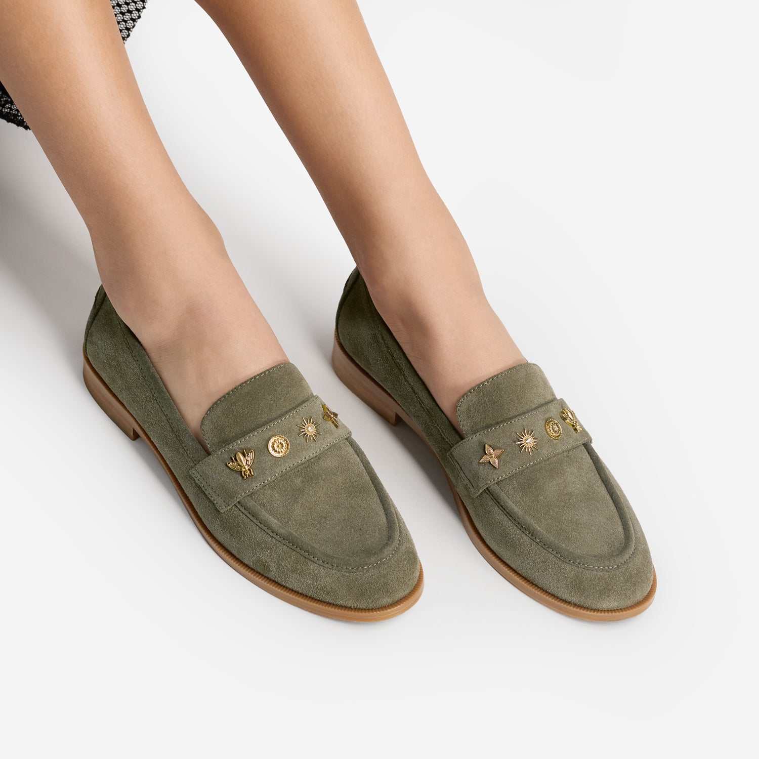 A pair of women's olive green suede almond toe loafers with gold-tone metal studs and star details on the vamp, shown on a model's feet against a light gray background.