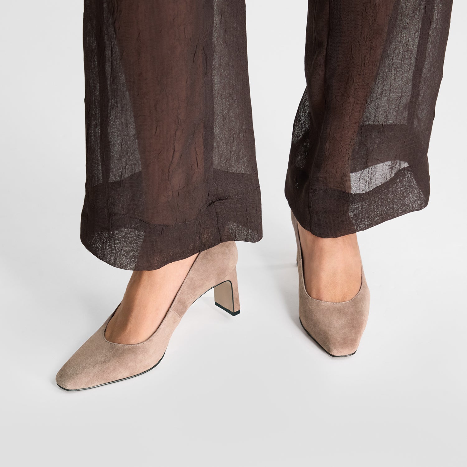 Beige suede medium-heeled shoes worn with sheer brown pants on a white background