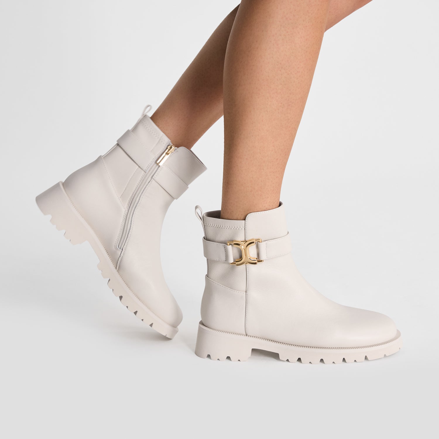 Flat ivory ankle boots with gold buckles worn by a person on a light gray background by Nicole Rebstock. 