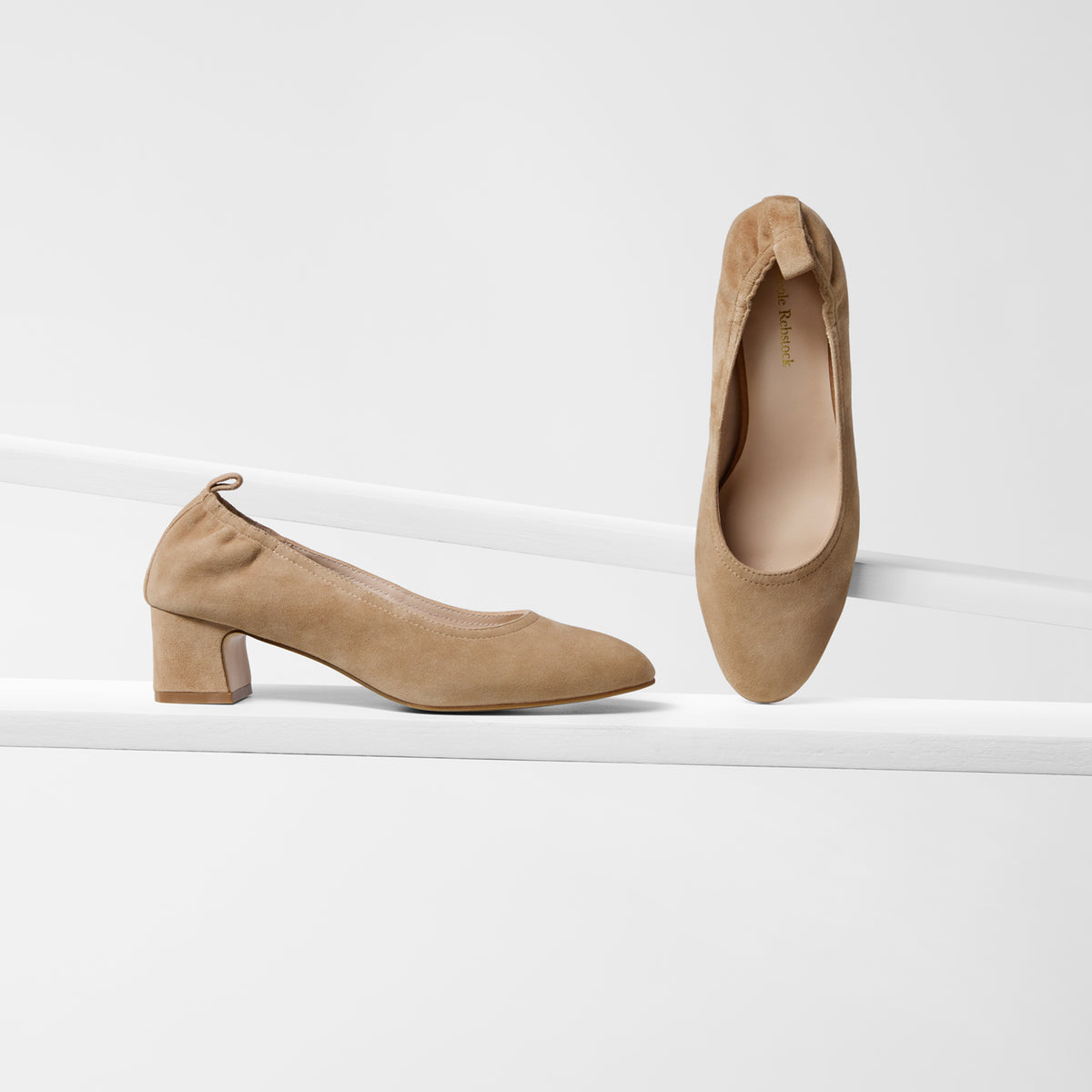 A pair of beige suede high-heeled pumps, one shown from the side profile and the other from the top view, displayed on a clean white geometric riser.