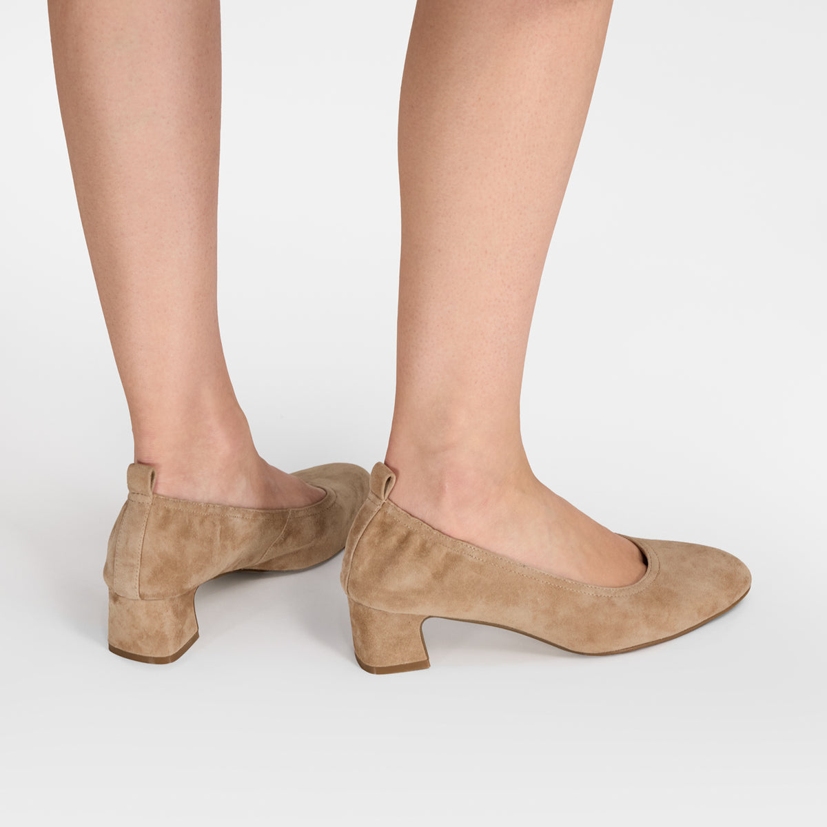 A pair of beige suede block-heel pumps shown on a model's feet from a rear three-quarter angle against a light gray studio background. Made by Nicole Rebstock.