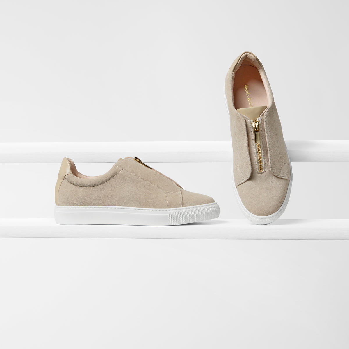 A pair of beige slip-on sneakers with white soles, one shown from the side and the other from the top, featuring a gold zipper on tongue detail on a clean white background.