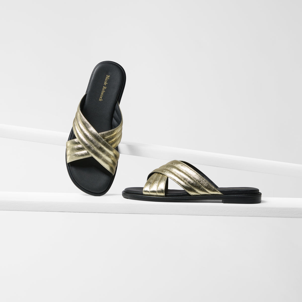 A pair of black slide sandals with metallic gold crisscross straps, displayed on a clean white geometric riser.