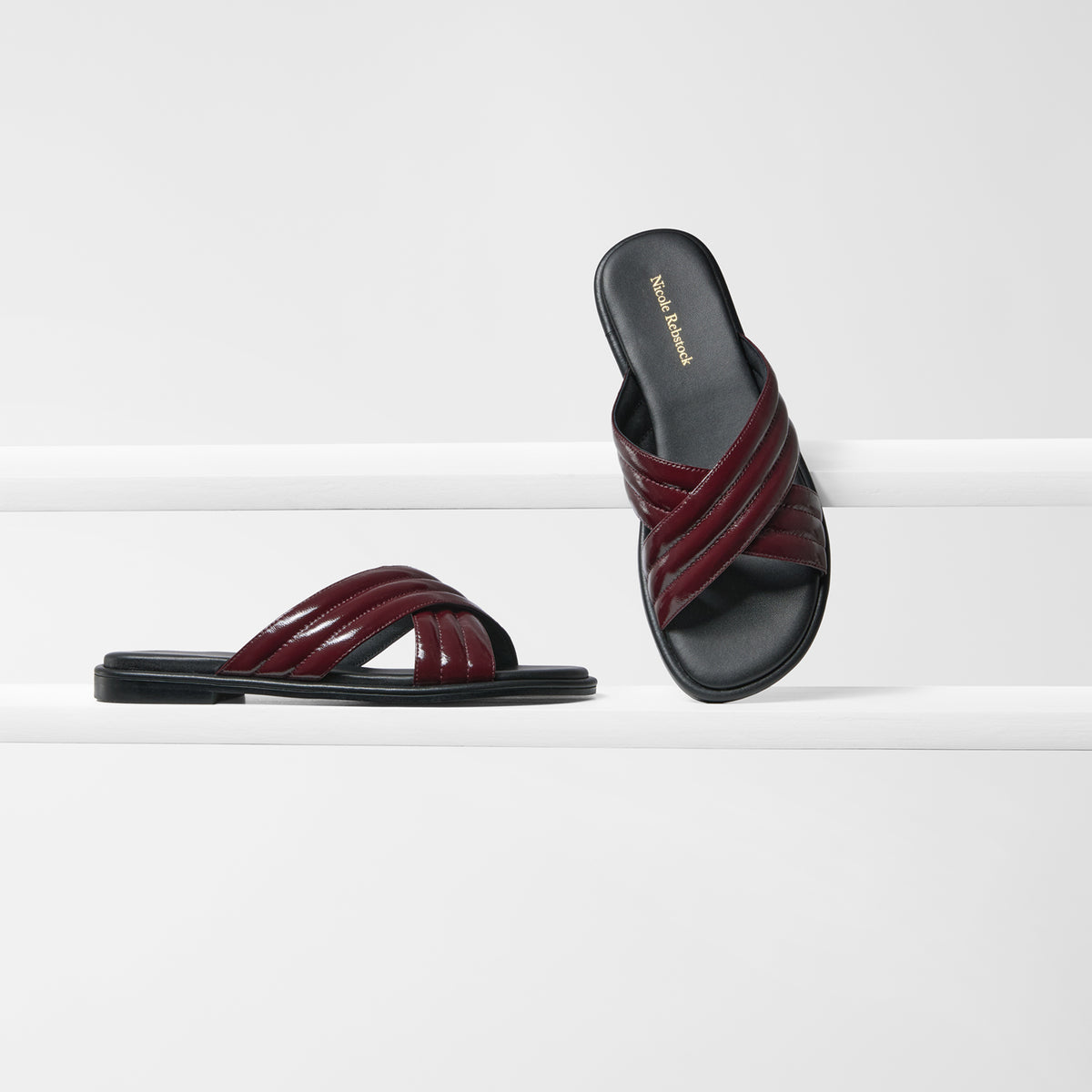 A pair of women's slide sandals with glossy burgundy leather crisscross straps and black soles, displayed on a clean white geometric riser.