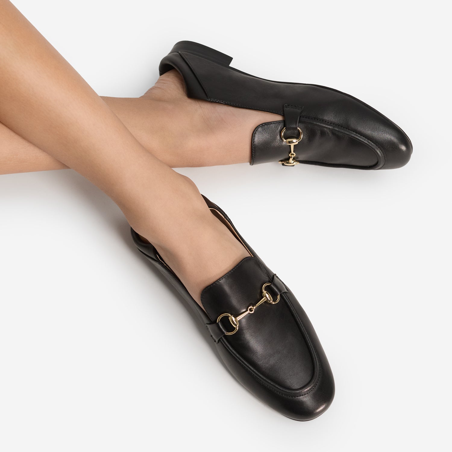 A pair of black leather loafers with gold-tone horsebit hardware, shown on a model's feet against a clean white background.