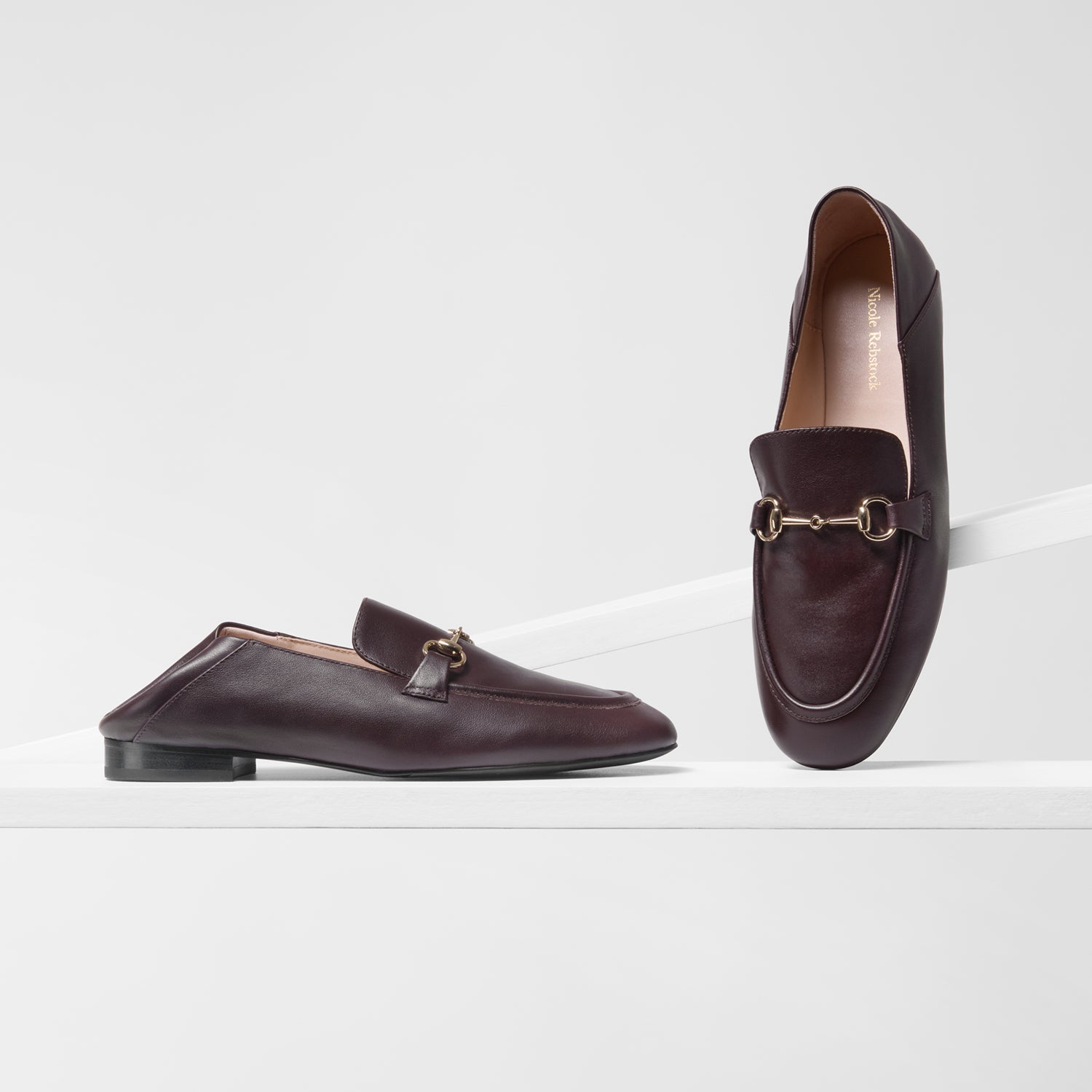 A pair of dark brown leather loafer mules with gold-tone horsebit detailing, displayed on a clean white geometric platform.