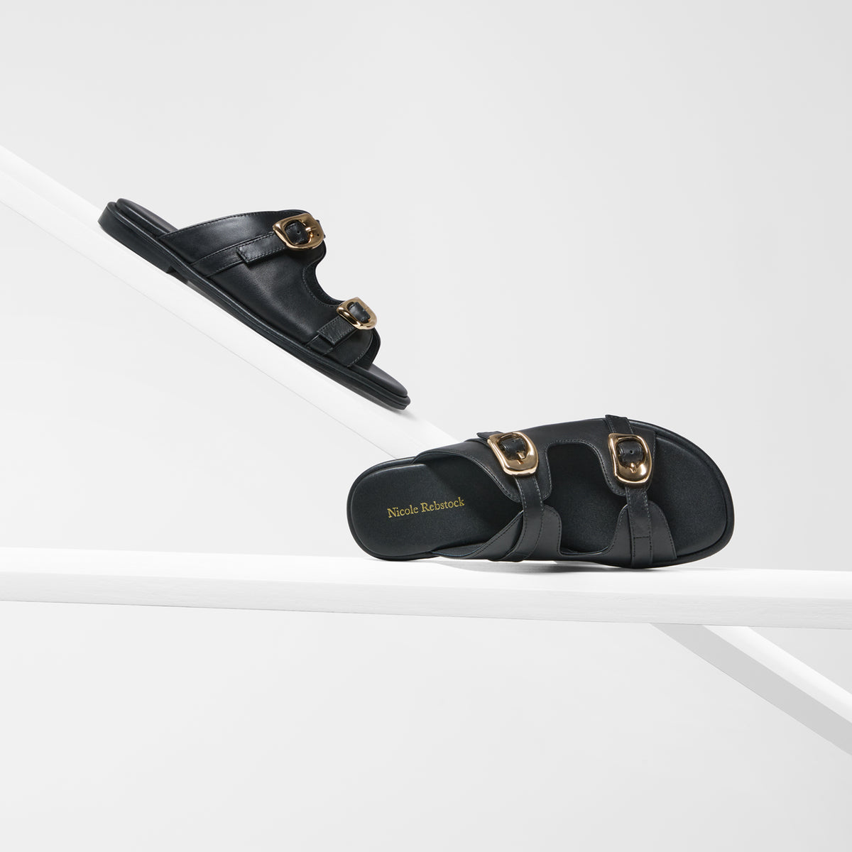 A pair of black double-strap slide sandals with gold-tone buckle details, displayed on a clean white geometric studio background. One sandal is shown from a side profile resting on a diagonal platform, and the other is positioned below it showing the insole branding.