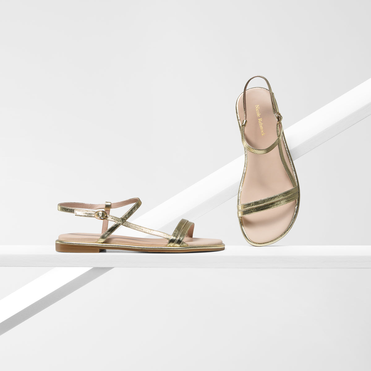 A pair of metallic gold flat sandals with strappy detailing, shown on a clean white geometric studio background. One sandal is positioned upright on a diagonal platform, and the other lies flat on a lower platform.