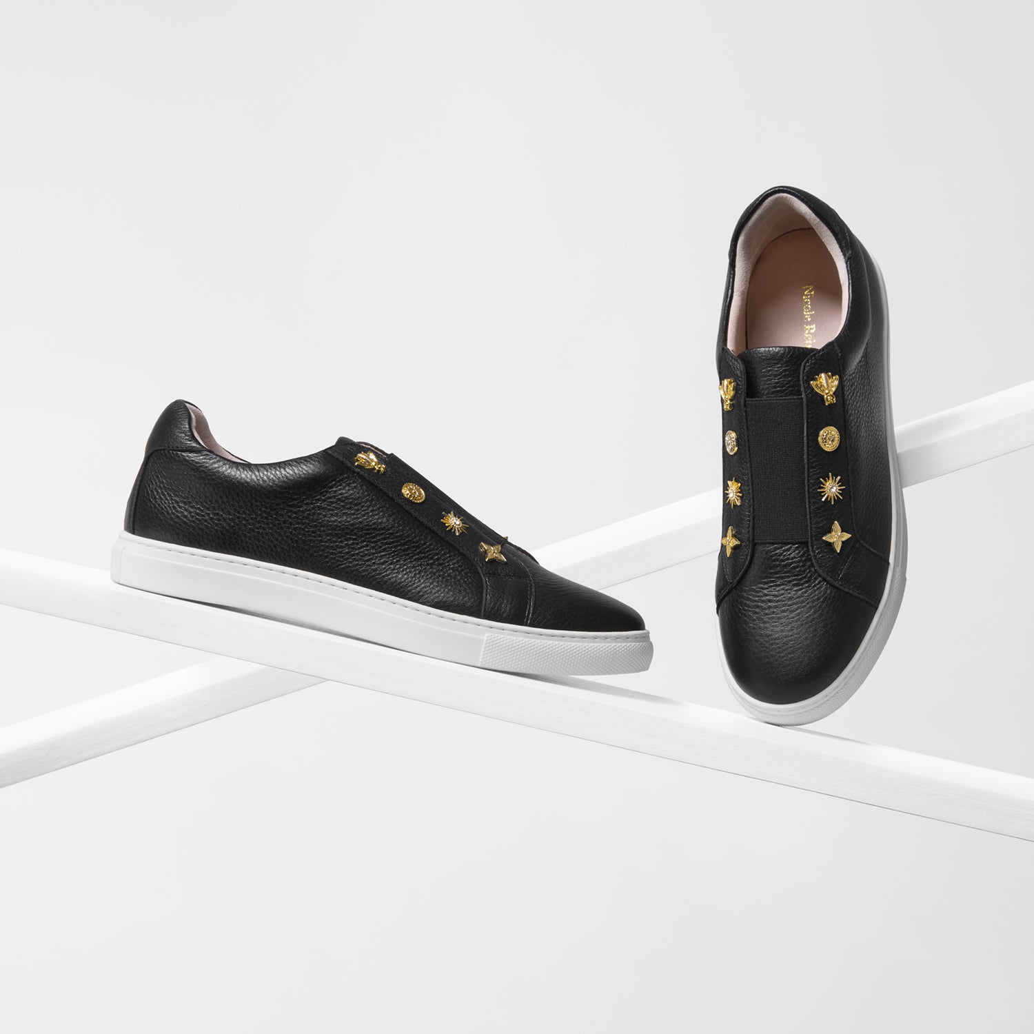 A pair of black leather slip-on sneakers by Nicole Rebstock with gold-tone decorative studs and star motifs, displayed on a clean white geometric platform.
