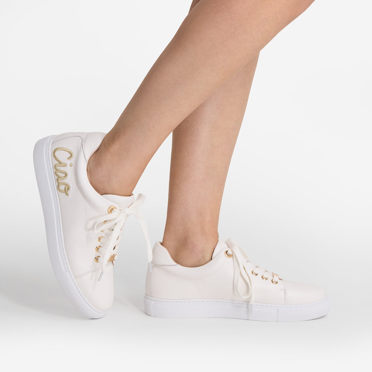 A pair of off-white leather low-top sneakers with gold accents, shown on a model's feet against a clean white studio background. The word 'Ciao' is printed in gold on the side of the left shoe.