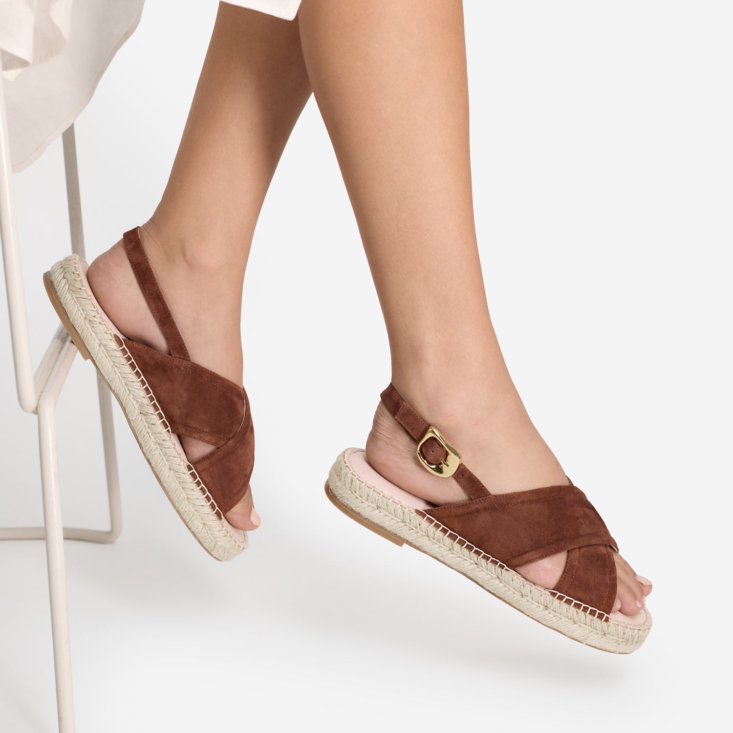A person wearing brown suede espadrille sandals with crisscross straps, ankle straps, and gold buckle details, shown against a clean white background.