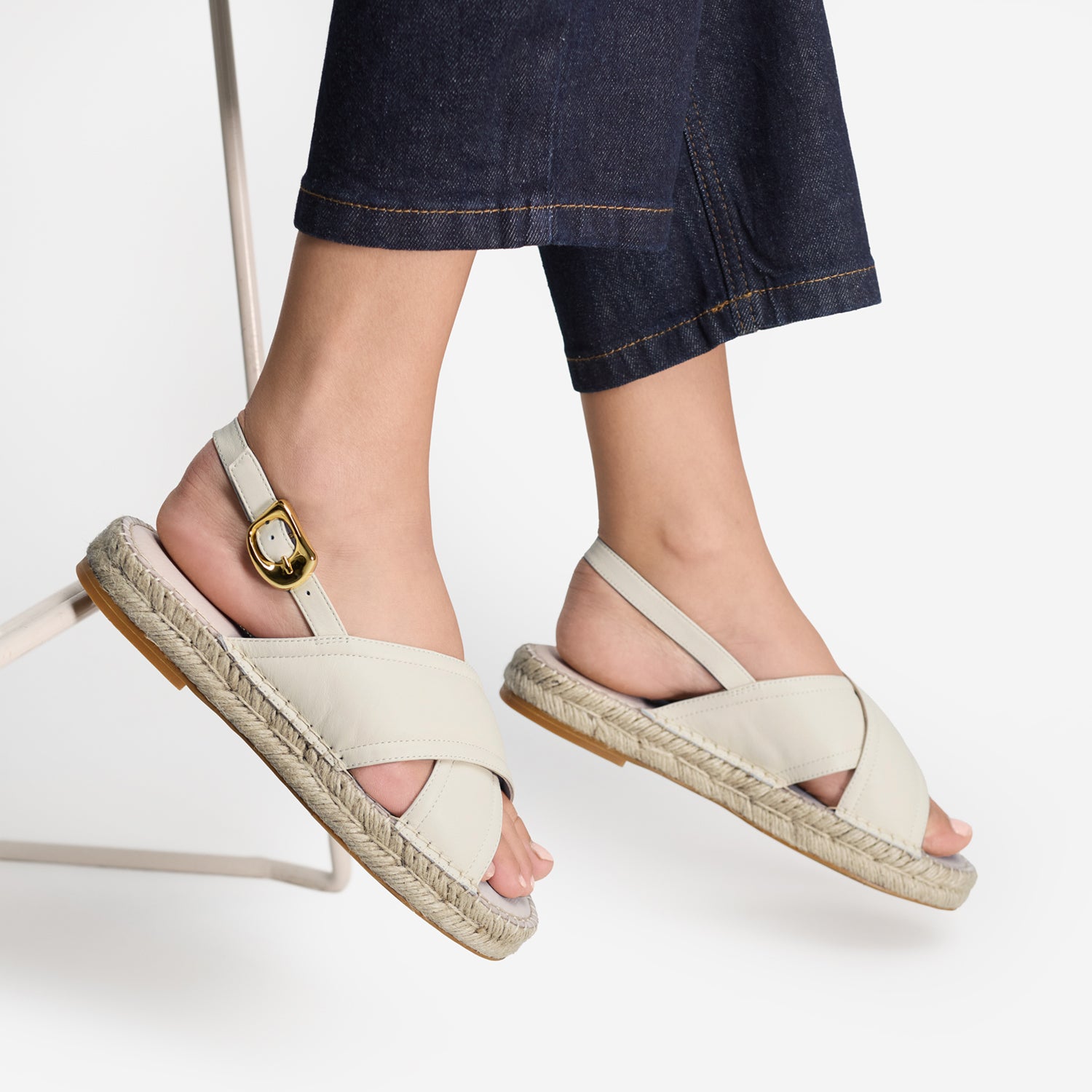 A pair of women's espadrille sandals with ivory leather straps, braided jute soles, and gold-tone buckle details, shown on a model wearing dark denim jeans against a clean white background.