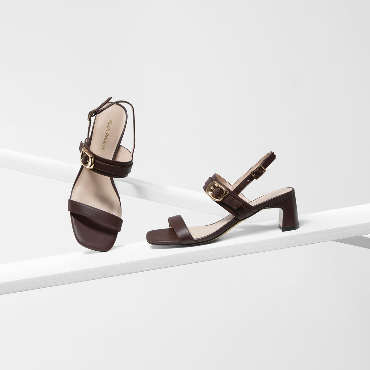 A pair of dark brown strappy sandals with block heels and gold-tone buckle details, displayed on a minimalist white geometric platform against a light gray background.