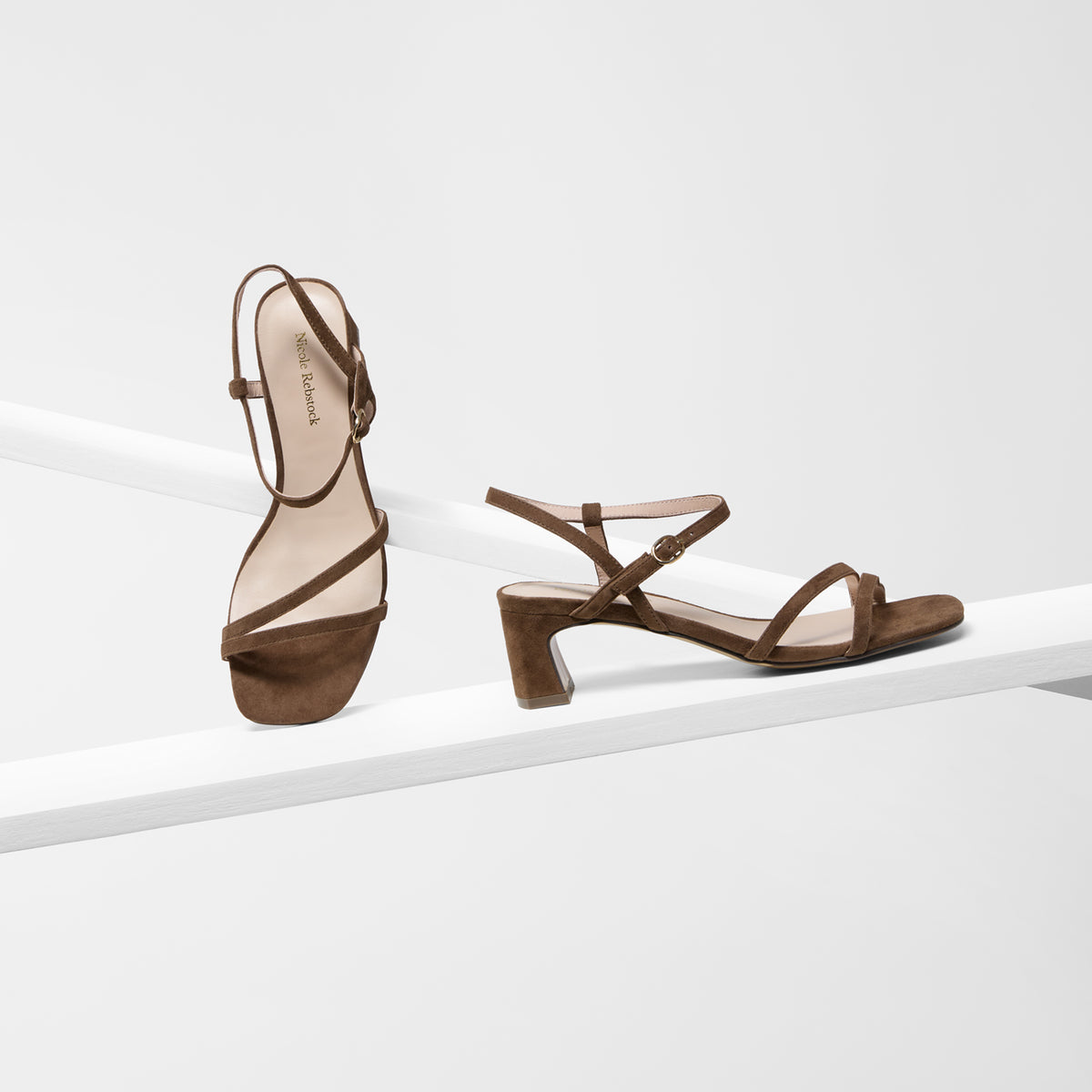 A pair of brown strappy sandal heels with block toes and low block heels, displayed on a minimalist white geometric platform against a light gray background.