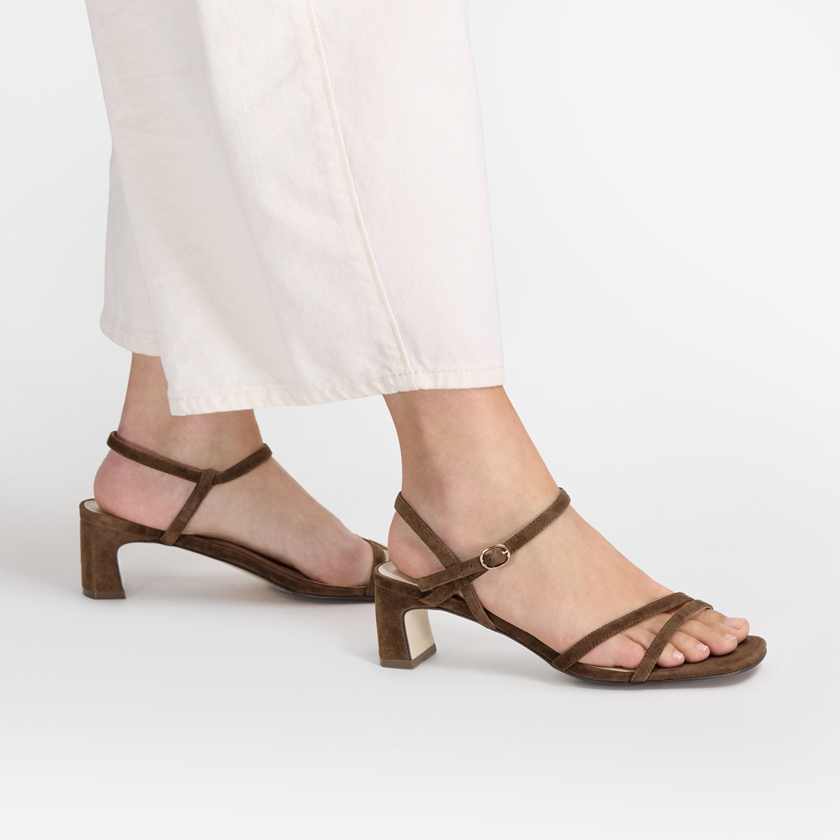 A pair of women's brown suede block-heel sandals with thin strappy details across the toes and a small buckle at the ankle by Nicole Rebstock, shown on a model wearing white pants against a light gray studio background.