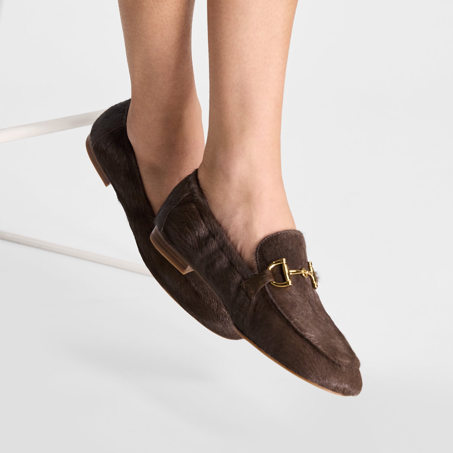 Brown loafers with a gold buckle on a light grey background worn by a model with elasticated back
