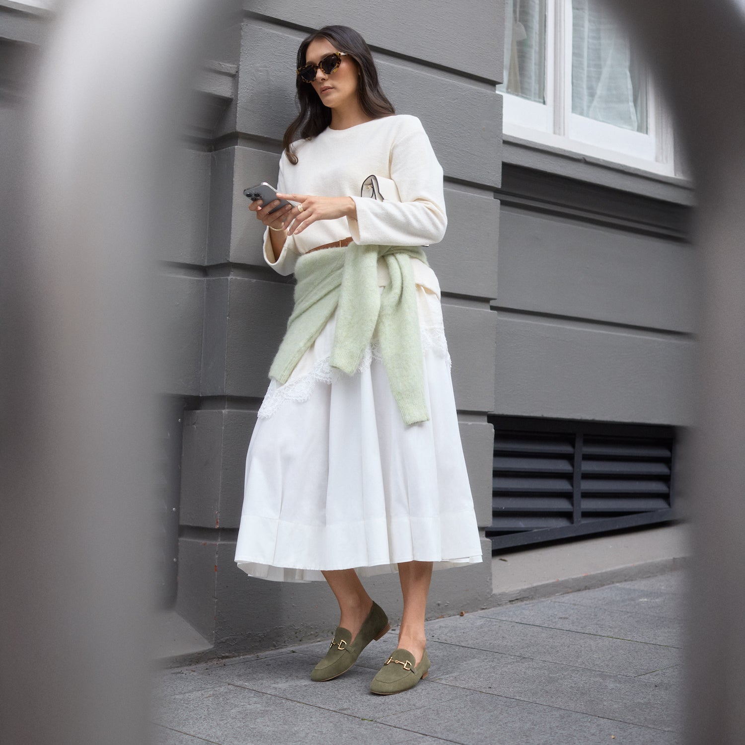 Woman in a white sweater and skirt wearing sage suede loafers.