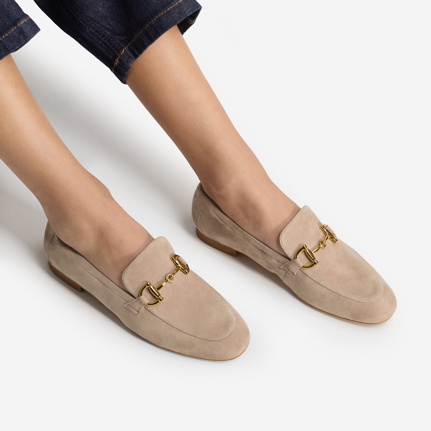 A pair of nude suede loafers  called Mel Loafer with gold horsebit hardware, shown on a model's feet with dark blue jeans cropped at the top, against a light gray studio background.