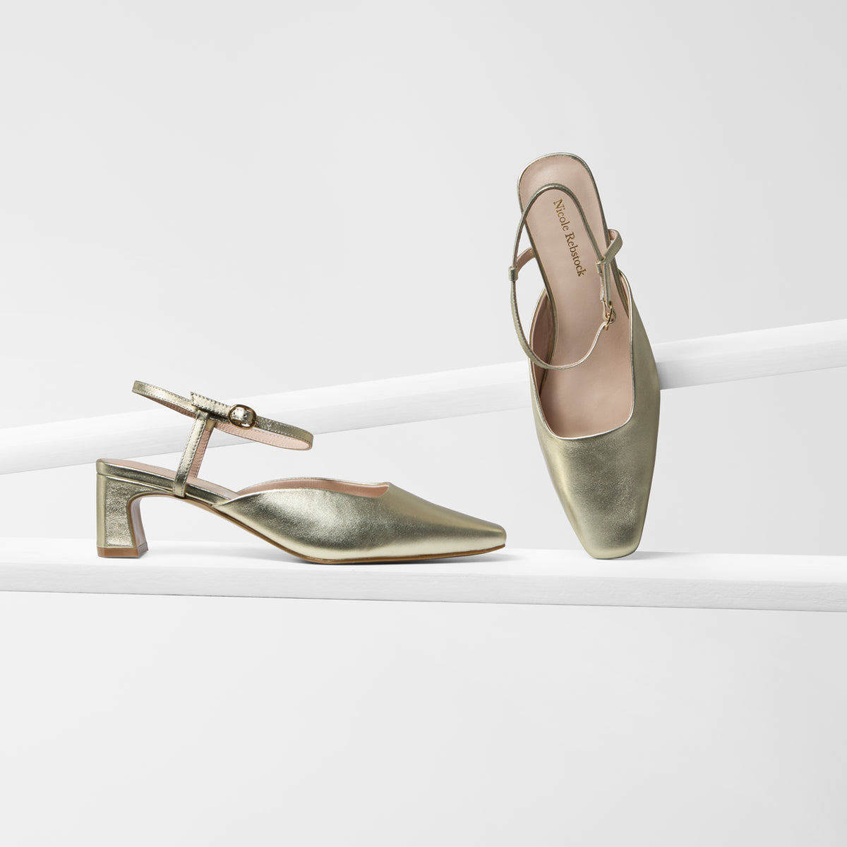 Two metallic gold slingback mule heels displayed on a clean white studio set. One shoe is shown in side profile on a white platform, and the other is angled to show the open toe and interior.