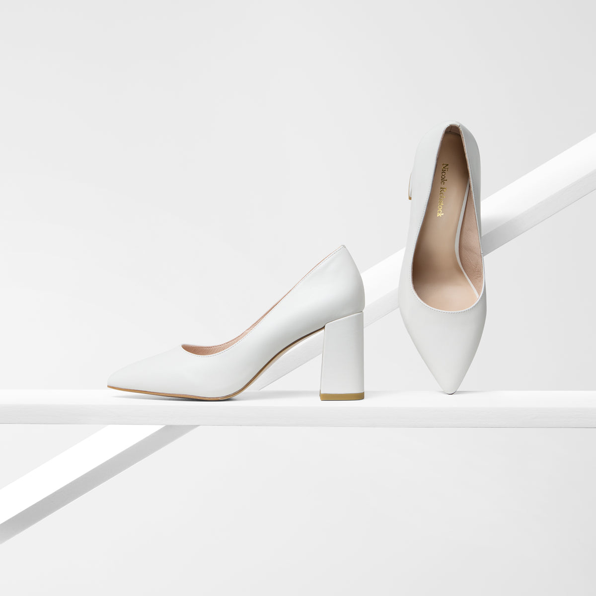 A pair of cream pointed-toe high heels with block heels, displayed on a minimalist white geometric platform. One shoe is shown in profile on the left, and the other is standing upright on the right, displaying the interior and the gold 'Mia Marigold' brand name on the insole.