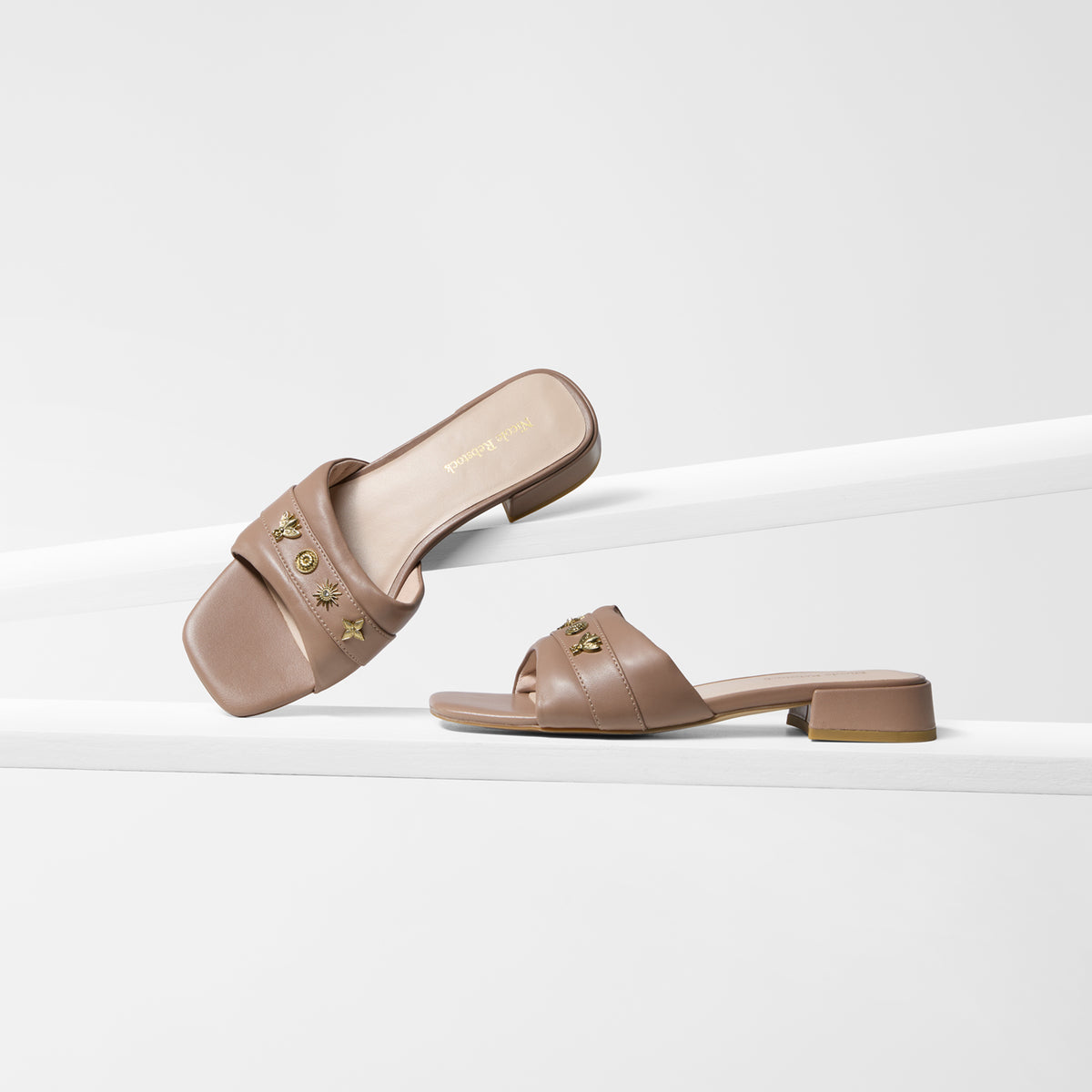 A pair of low heel taupe leather slide sandals with gold-tone studs, displayed on a clean white geometric riser.