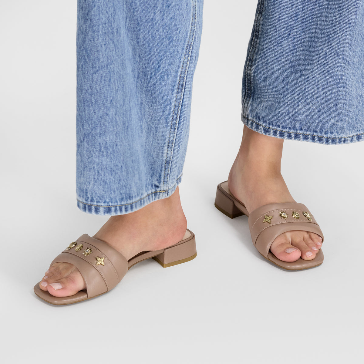 A person wearing flat taupe leather slide sandals with gold star embellishments and block heels by Nicole Rebstock, paired with light blue jeans, shown from the knees down on a light gray studio background.