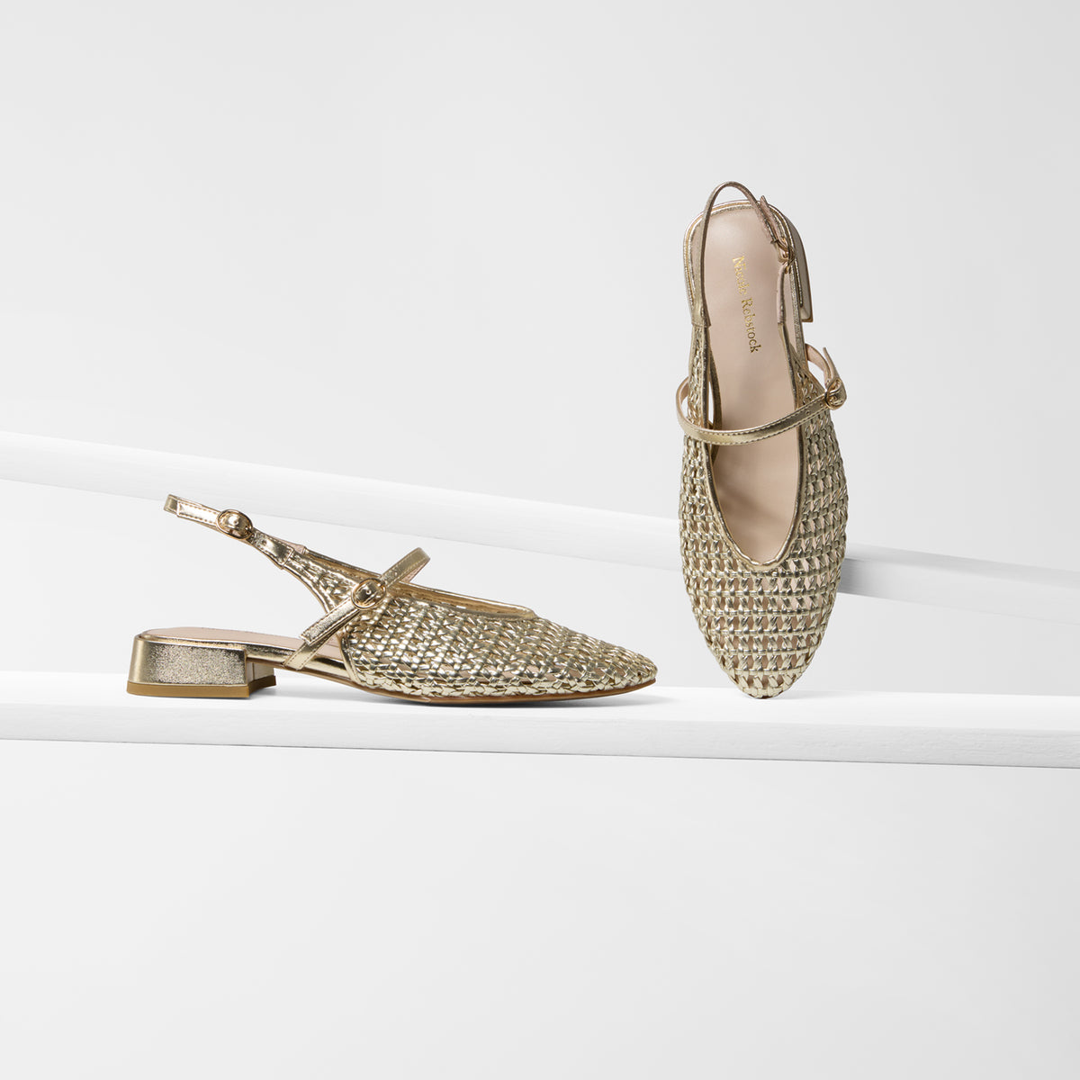 A pair of metallic gold slingback flat sandals with a woven textured upper, shown on a clean white platform against a white background. One sandal is positioned horizontally on the left, and the other stands vertically on the right.