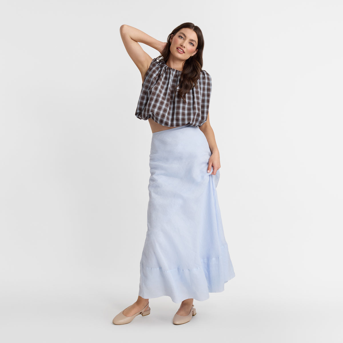 A woman modeling a light blue midi skirt paired with a brown and white plaid crop top and Nicole Rebstock's Sage Slingback, standing against a plain light gray studio background.