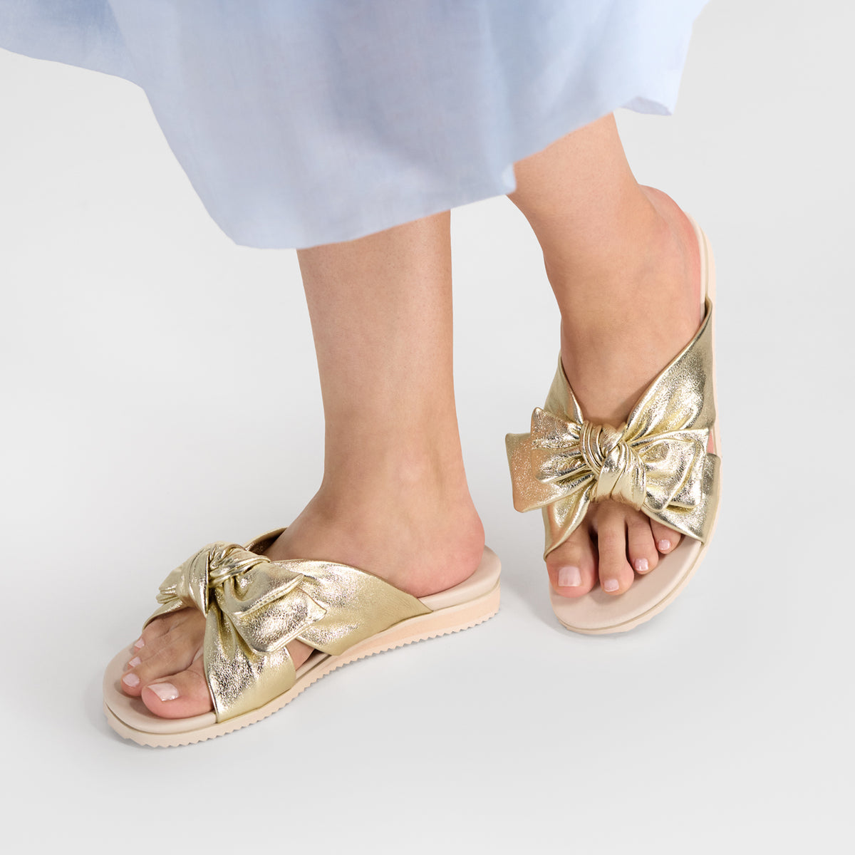 A pair of metallic gold slide sandals with large bow details called Salma Sandal by Nicole Rebstock, shown on a model's feet against a light gray studio background.