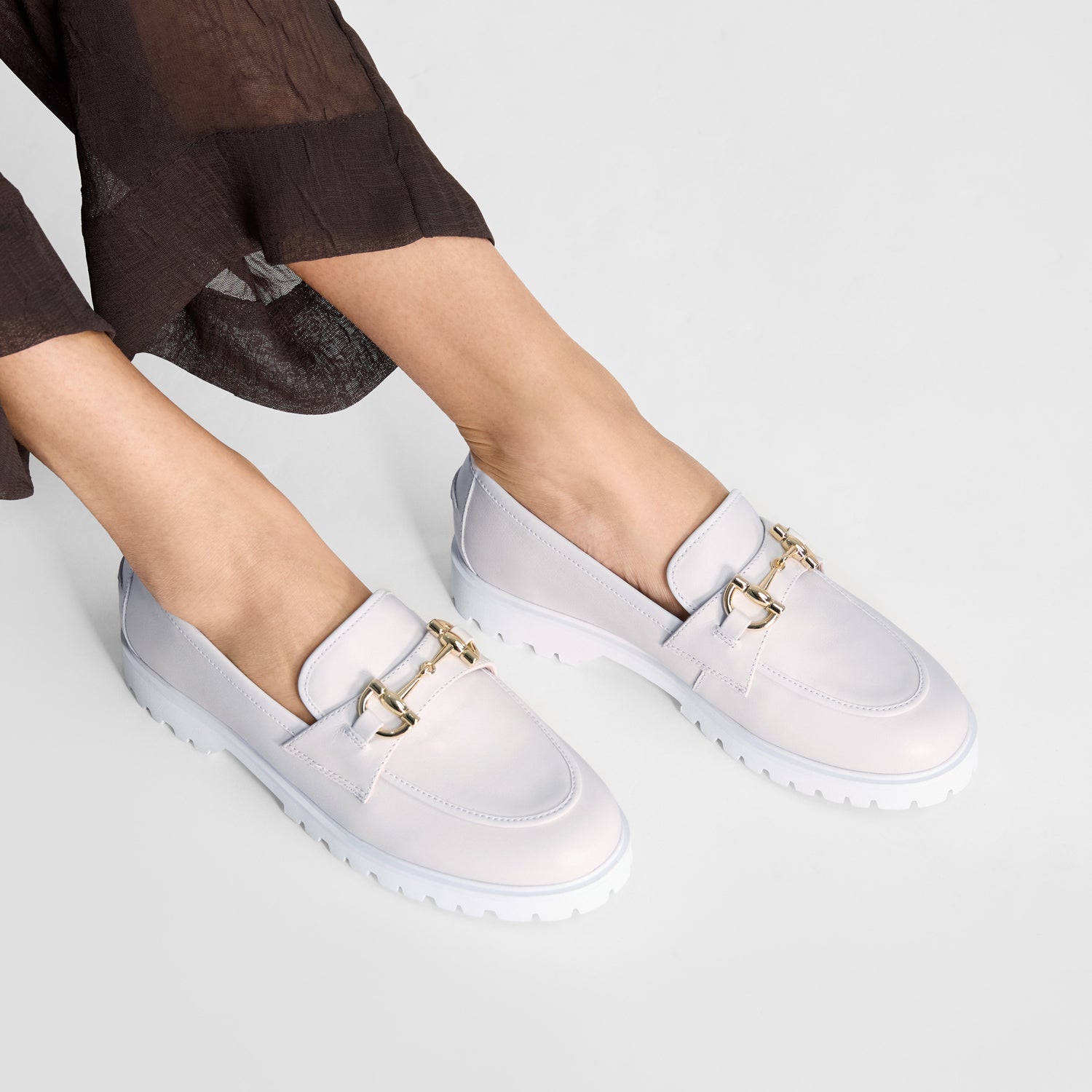 White loafers with gold accents and white tread worn by a person on a light grey background. 