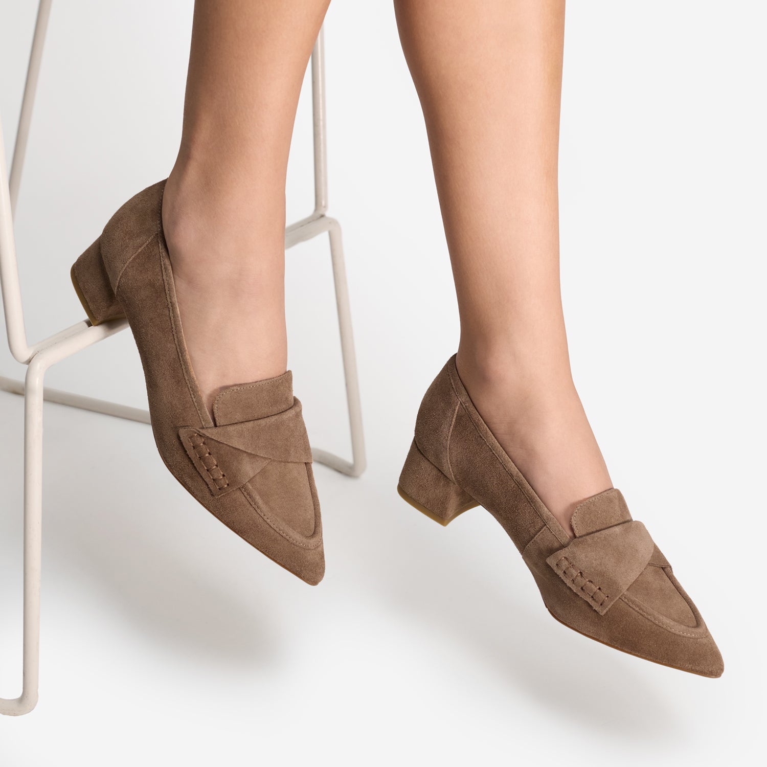 A pair of women's pointed-toe loafers in taupe brown suede, shown on a model's feet while sitting on a white metal stool against a light gray background.