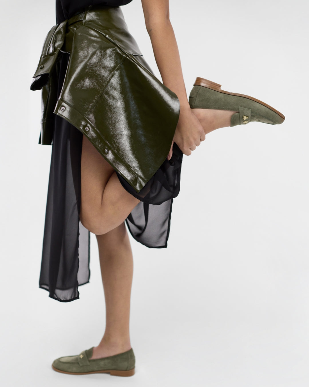A model wearing an asymmetrical high-low skirt with a glossy olive green patent leather outer layer and a sheer black underlayer, paired with olive green suede loafers, against a plain white studio background.