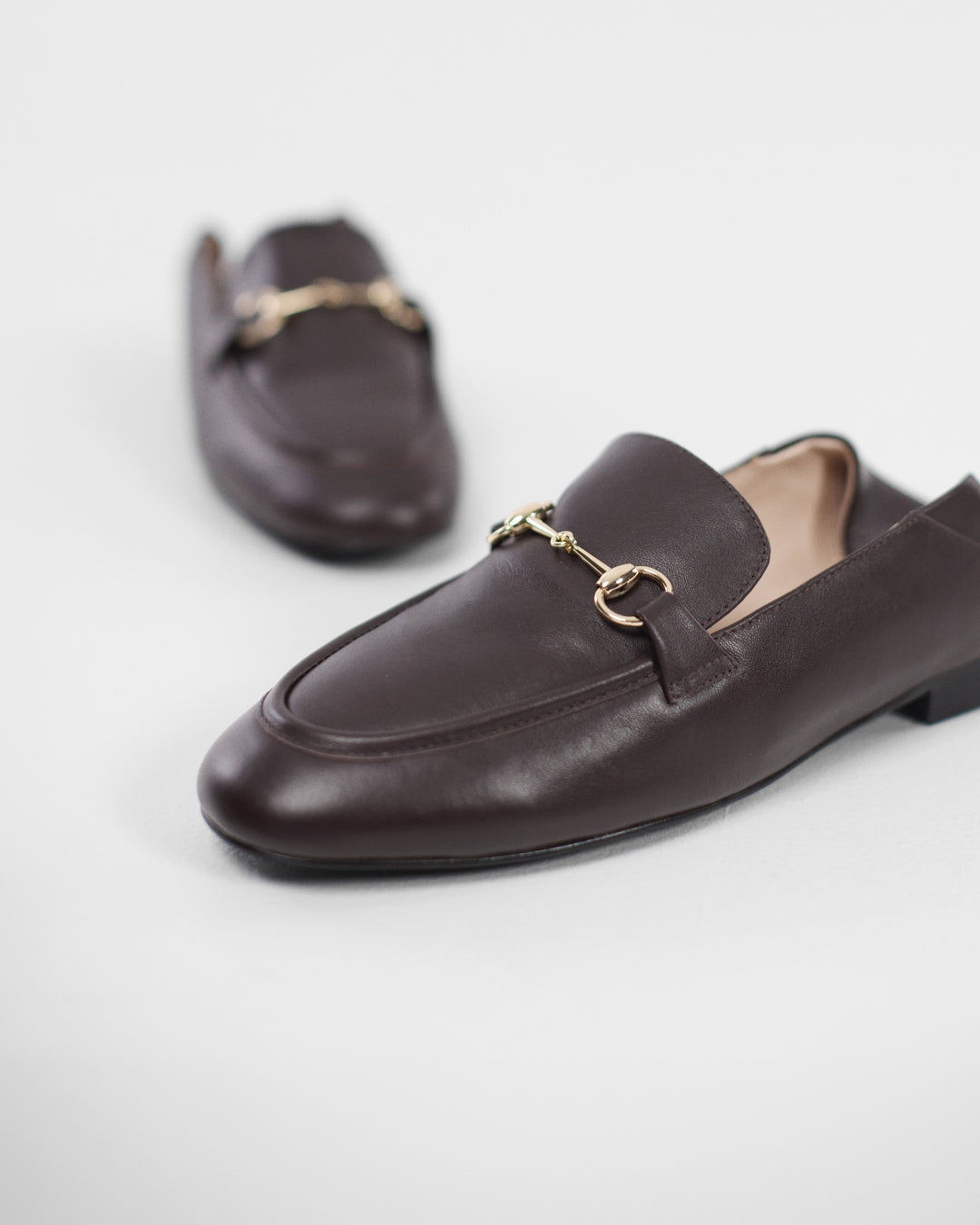 A pair of dark brown leather loafers with gold-tone horsebit hardware, shown on a clean light gray background.