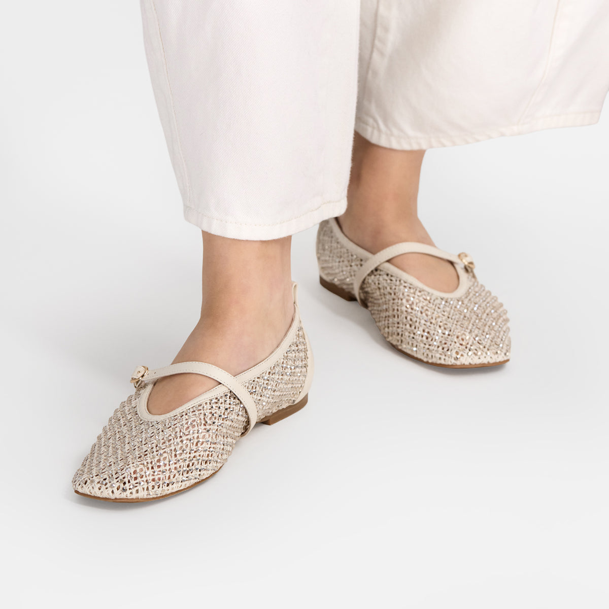 A pair of beige woven Mary Jane flats with a single leather strap and a small gold-tone bow, shown on a model wearing white pants against a light gray studio background.